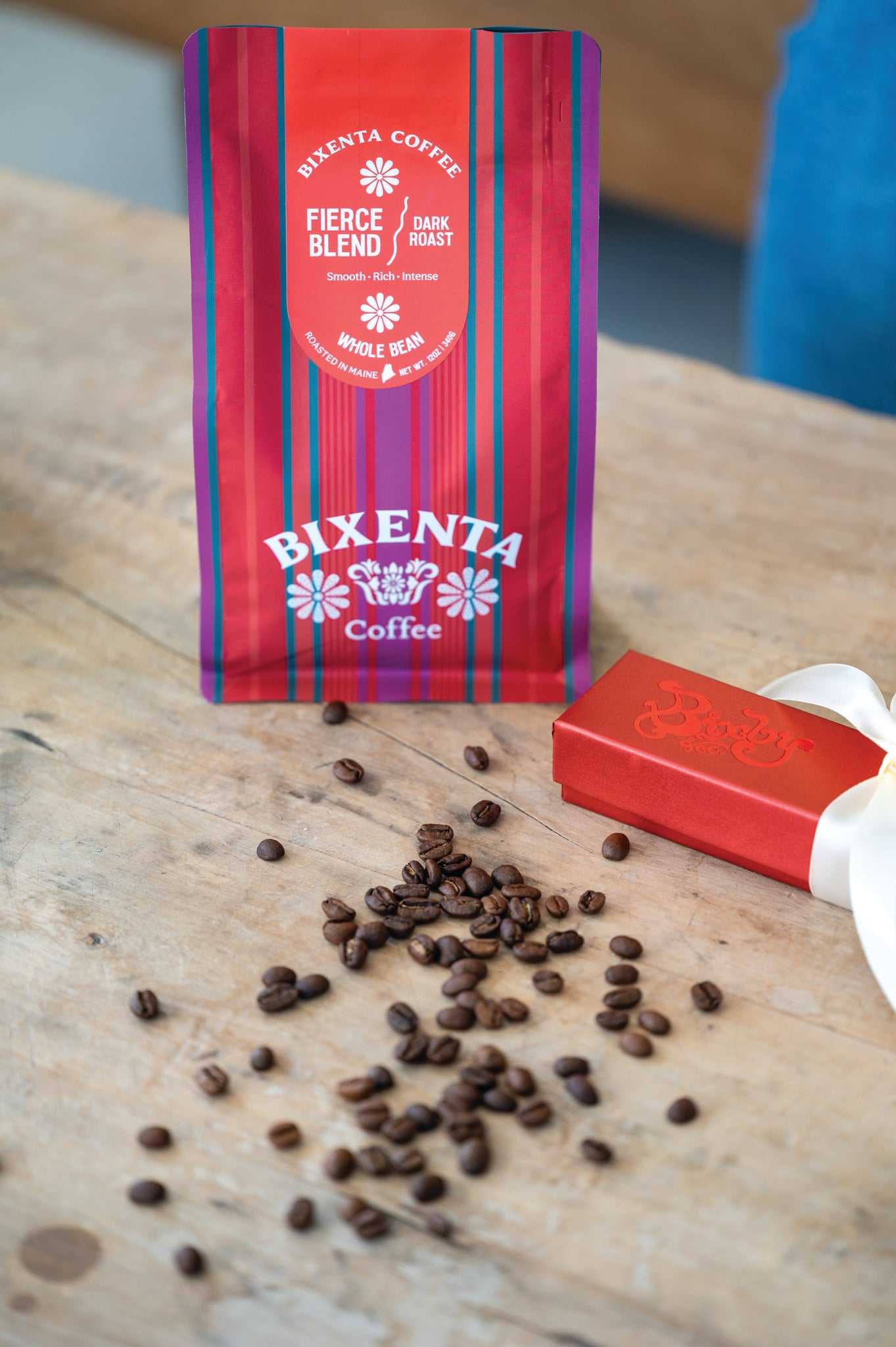 A red bag of Bixenta Coffee Fierce Blend dark roast whole beans sits on a wooden table next to scattered coffee beans and a small red gift box with a white ribbon.