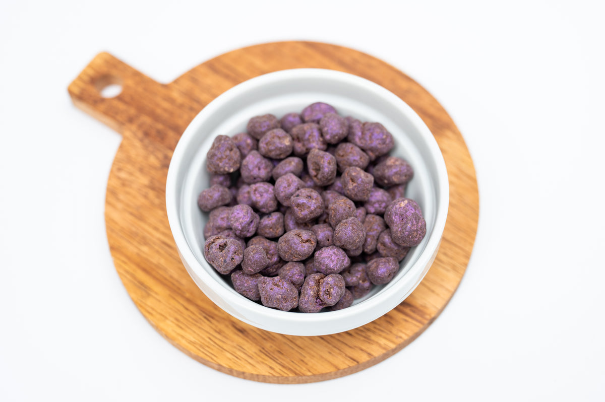A white ceramic bowl filled with Bixby Chocolate Dark Chocolate Covered Blueberries sits on a circular wooden board against a white background.