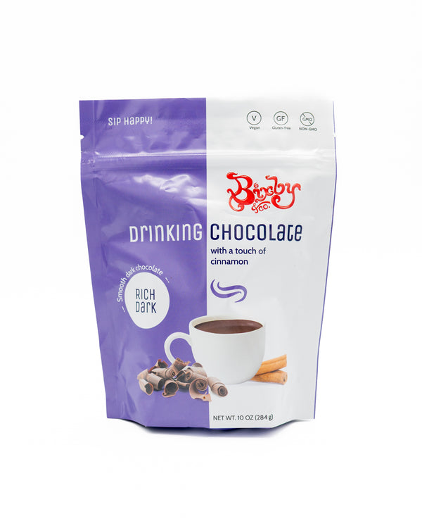 A purple and white bag of Bixby Chocolates' Non GMO Drinking Chocolate Mix, featuring a cup of vegan hot chocolate, chocolate curls, and cinnamon sticks. This 10 oz dark chocolate mix boasts a bold cinnamon flavor.