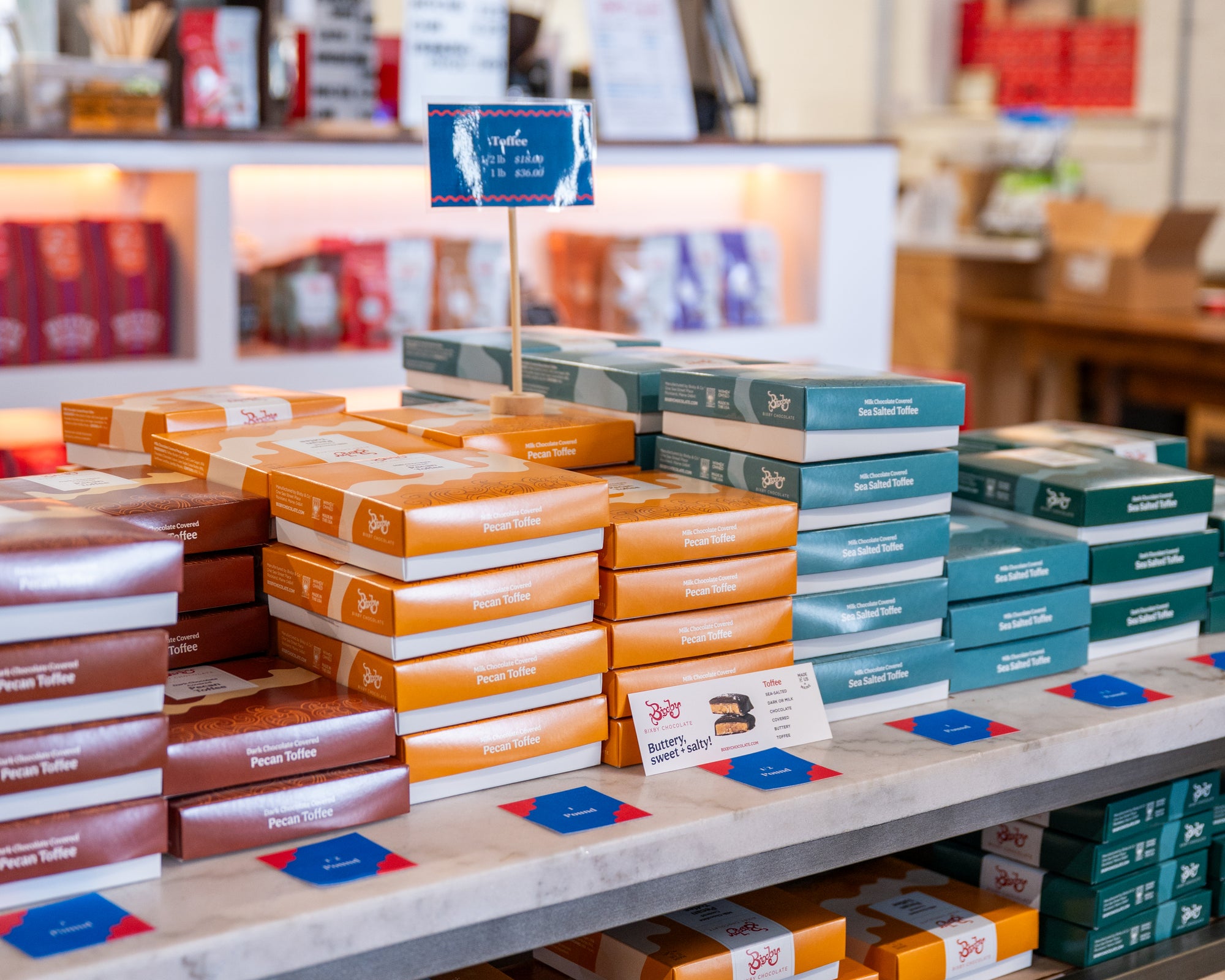 Stacks of colorful boxes of pecan toffee are displayed on a store table, with small signs and price cards. Shelves with more products and a counter are visible in the background.