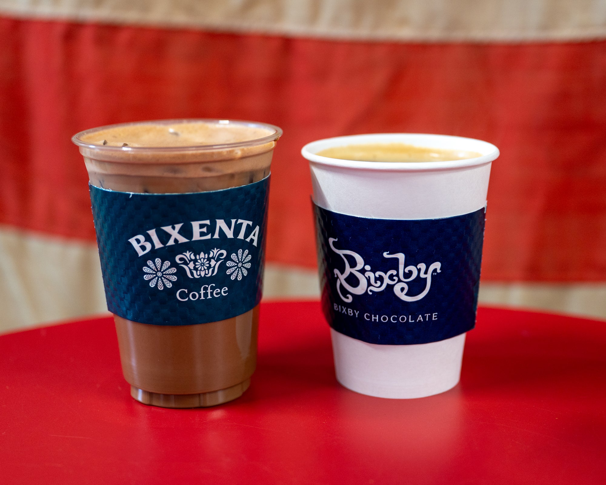 Two takeaway drinks sit on a red table: an iced coffee in a plastic cup labeled Bixenta Coffee and a hot drink in a paper cup labeled Bixby Bixby Chocolate, with a red and beige background.