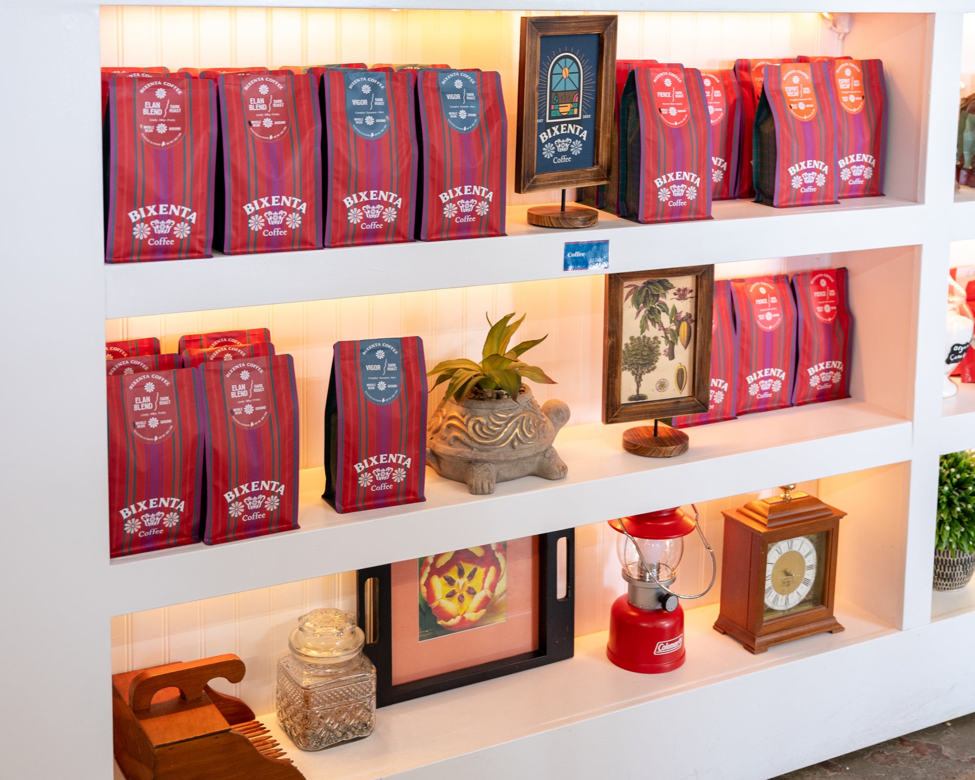 A white shelving unit displays multiple red bags of Bixenta coffee, decorative plants, a framed picture, a red lantern, a clock, and a jar filled with what appears to be coffee beans.