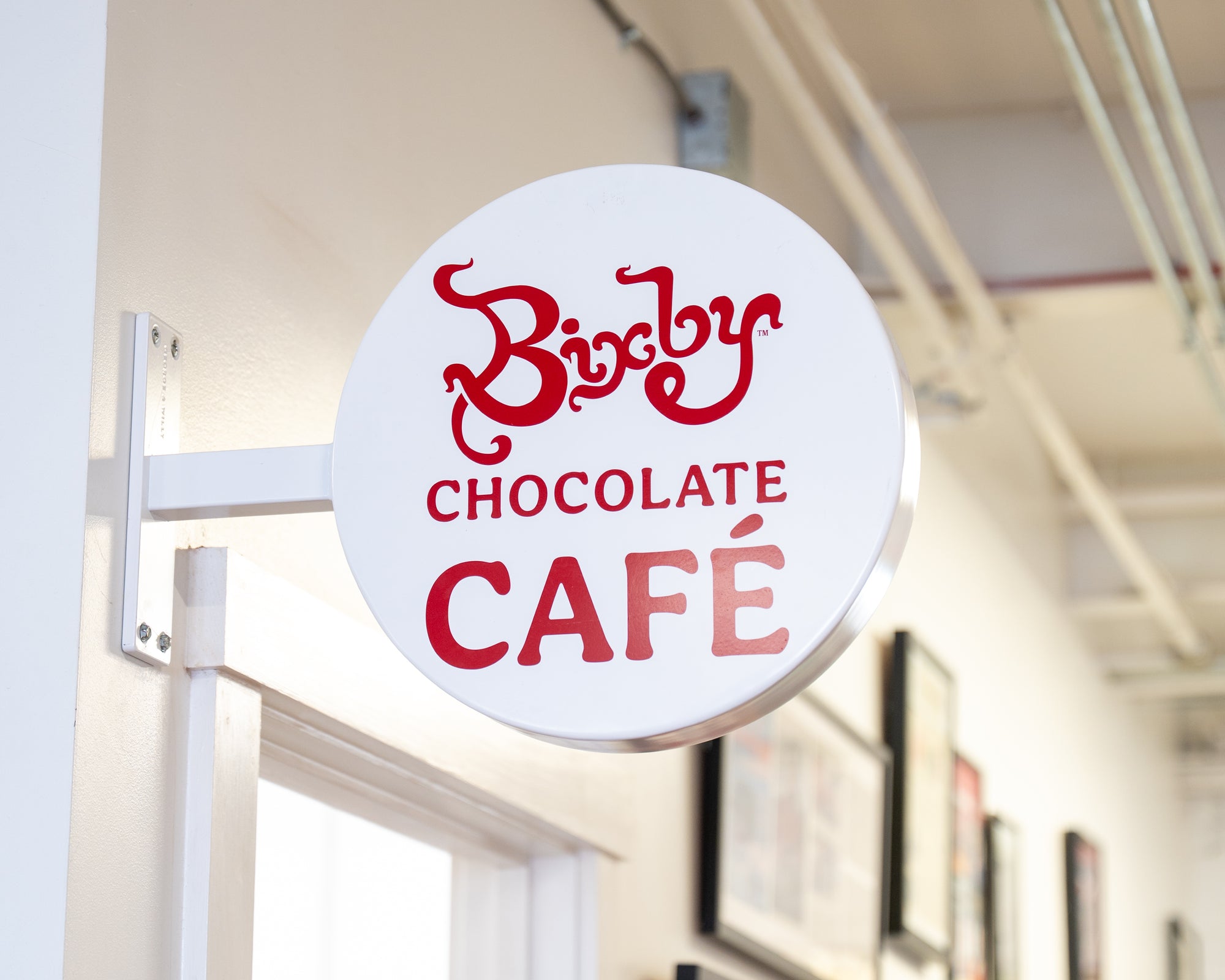 A round white sign with red text reading Bixby Chocolate Café is mounted on a wall inside a building with white walls and visible ceiling pipes.