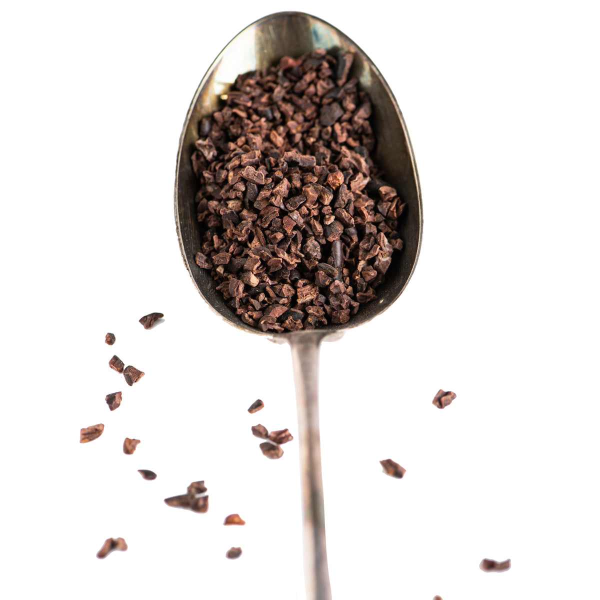 A vintage spoon filled with Bixby Chocolates Organic Dominican Republic Cacao Nibs—celebrated superfood—with some nibs scattered around, isolated on a white background.