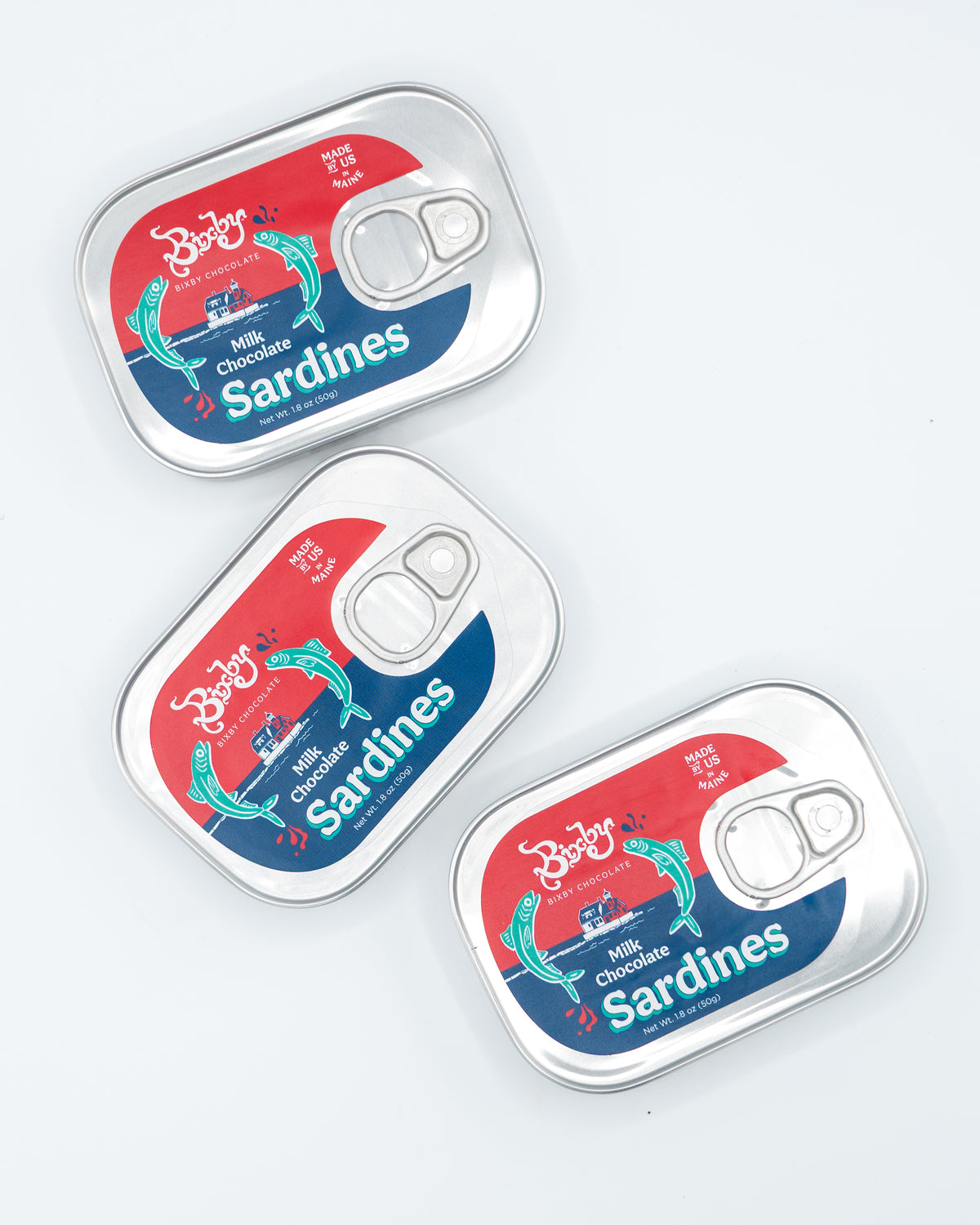 Three silver tins of Bixby Chocolates’ Chocolate Sardines, each with colorful fish-themed labels, are arranged on a white surface. These whimsical treats resemble sardine cans and make a unique Maine souvenir.