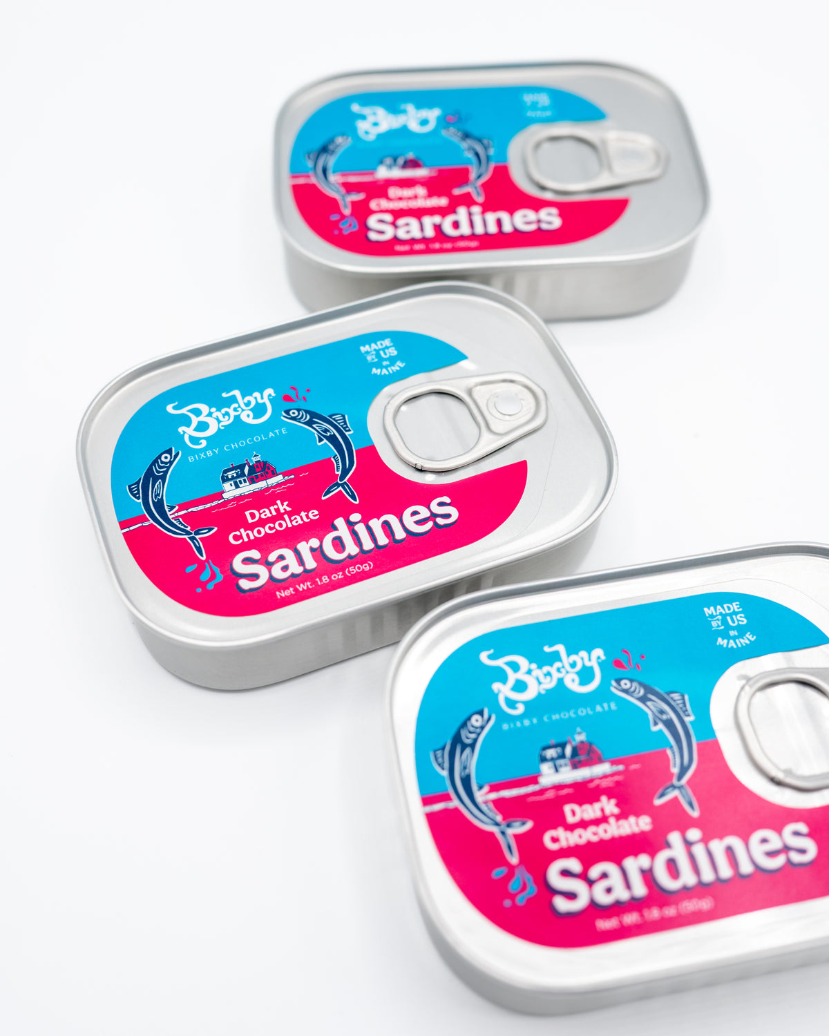 Three tins of Bixby Chocolates’ Chocolate Sardines, featuring bright blue and pink labels with a fish illustration and bold white text, are arranged on a white surface—a perfect Maine souvenir.