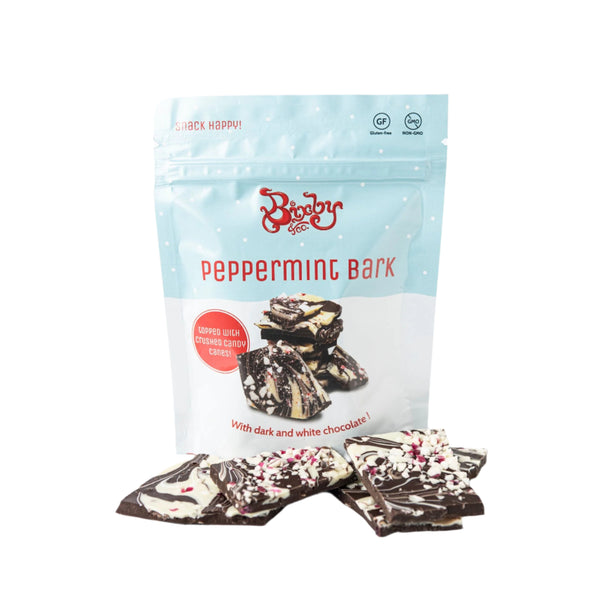 A bag labeled Bixby Chocolates Peppermint Bark sits behind pieces of artisan chocolate bark topped with crushed candy canes. The festive packaging makes it an ideal holiday gift.