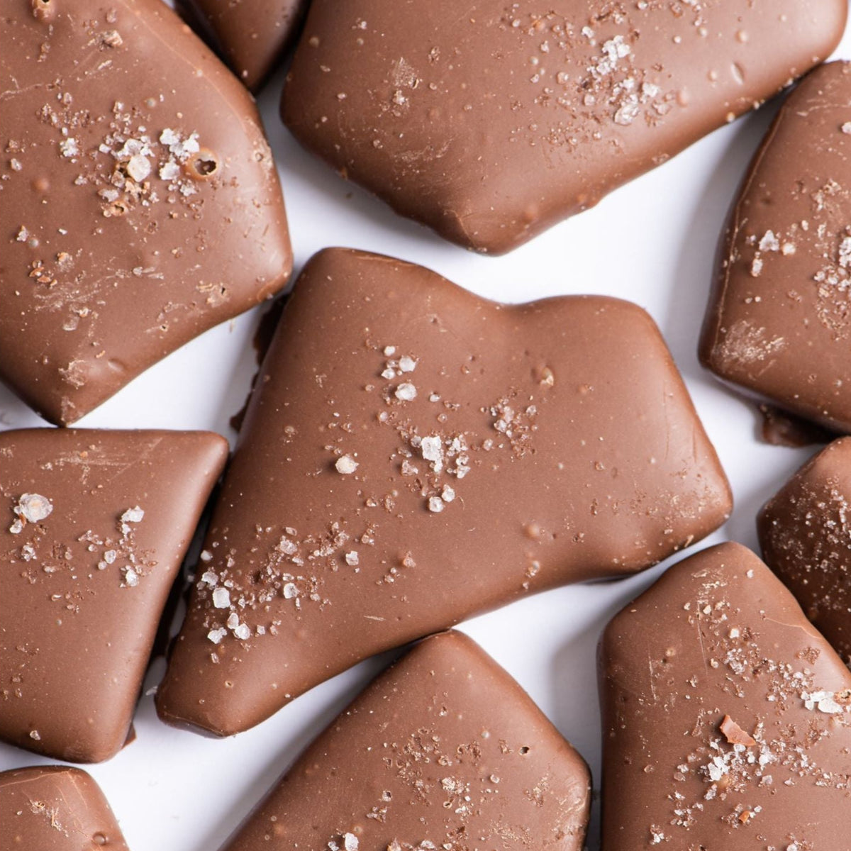 Bixby Chocolates Milk Chocolate Toffee features buttery toffee pieces coated in creamy milk chocolate and dusted with coarse sea salt, all beautifully arranged on a white surface.