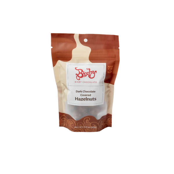 A brown and beige resealable pouch of Bixby Chocolates Dark Chocolate Hazelnuts stands upright against a white background, featuring swirly designs, the Bixby Chocolates logo, and highlighting its roasted hazelnut pieces inside.