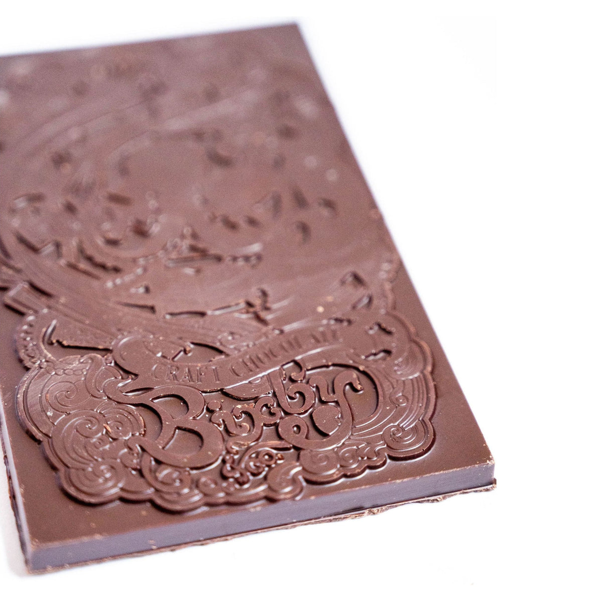A close-up of a rectangular Organic Milk Chocolate Maine Sea Salt Bar by Bixby Chocolates, featuring intricate decorative engravings and branding on its surface, set against a white background.