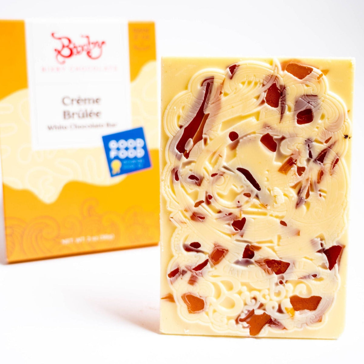 A white chocolate bar with caramel swirls and shards, made with organic ingredients and real vanilla bean, stands before its yellow-and-white box labeled Organic Crème Brûlée White Chocolate Bar by Bixby Chocolates, featuring a Good Food Awards sticker.