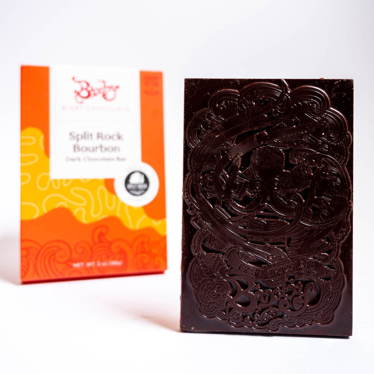 A dark chocolate bar with an intricate embossed design stands upright before an orange and white box labeled “Organic Split Rock Bourbon Bar” by Bixby Chocolates, crafted from organic single-origin cacao for a rich, distinctive flavor.