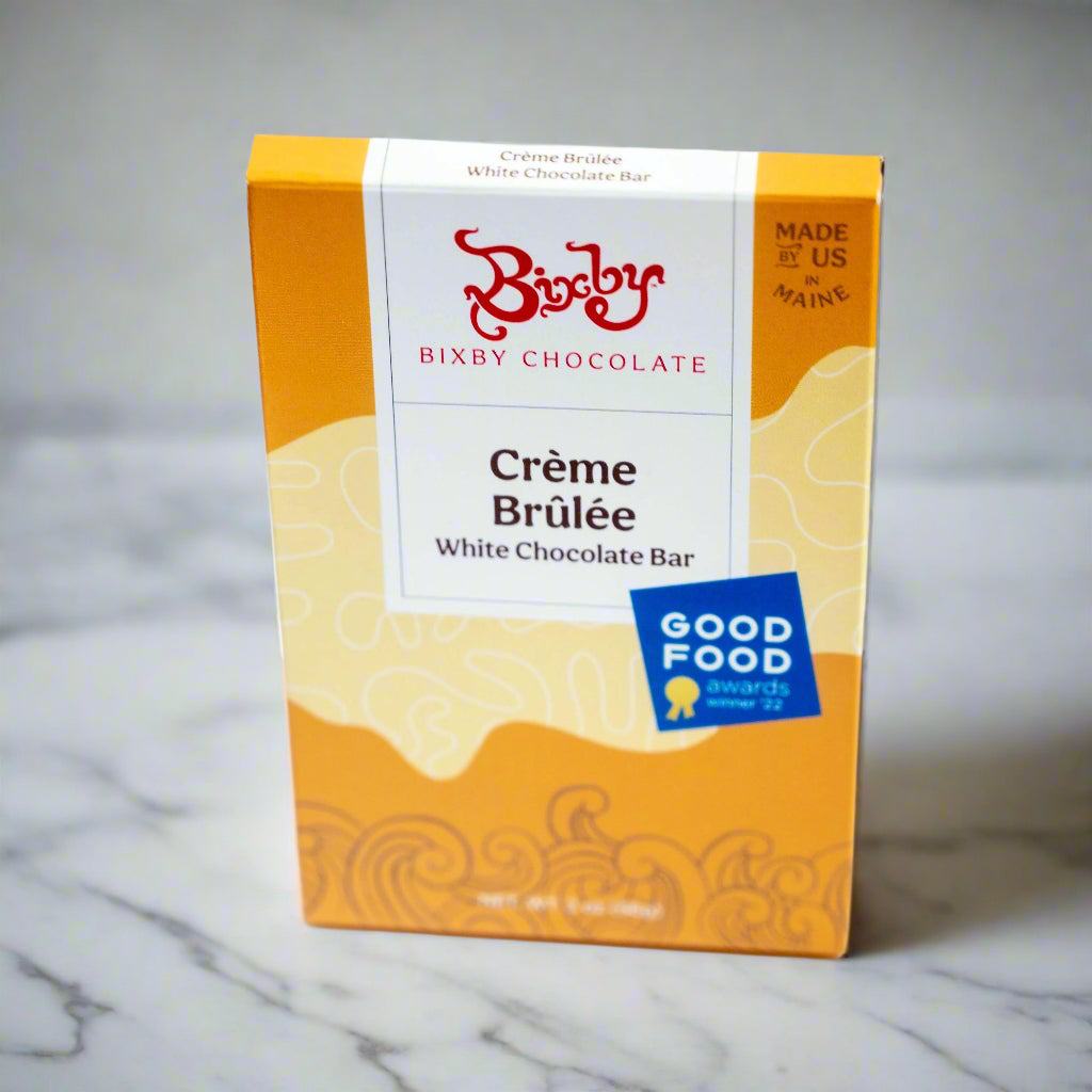 A box of Bixby Chocolates Organic Crème Brûlée White Chocolate Bar, made with organic ingredients and vanilla bean, sits on a marble surface. The yellow and white packaging displays a Good Food Awards 2022 sticker and “Made in Maine” text.