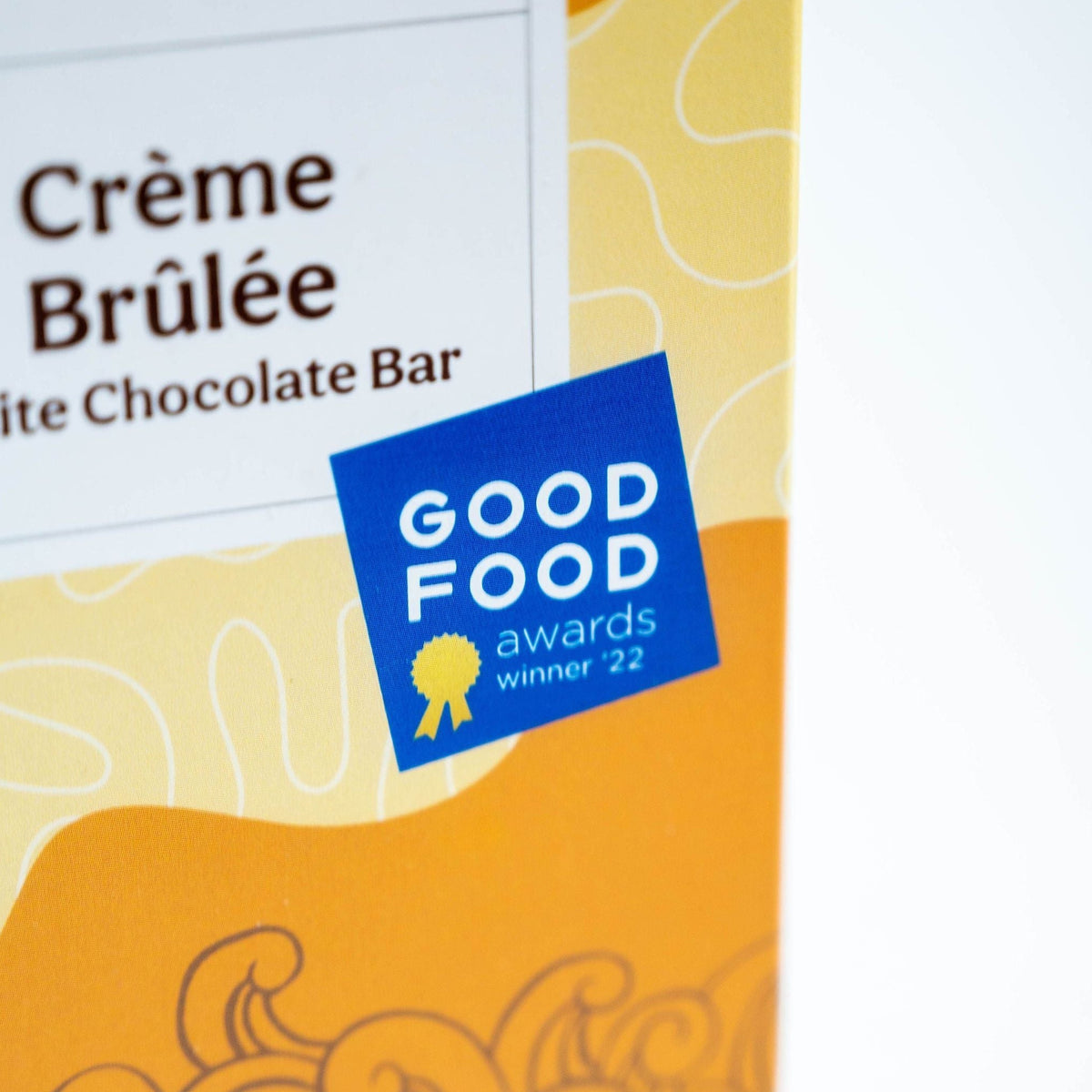 Close-up of a Bixby Chocolates Organic Crème Brûlée White Chocolate Bar package with aromatic vanilla bean and a blue Good Food Awards '22 sticker on a yellow and white patterned background.