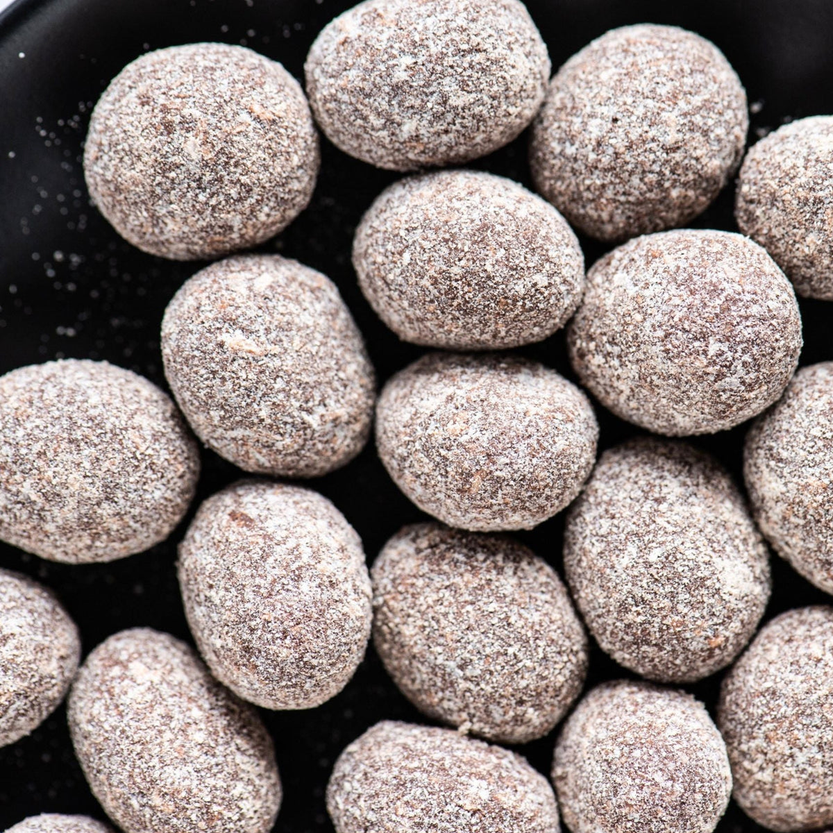 A close-up of Bixby Chocolates' Dark Chocolate Coconut Almonds, lightly dusted with powdered sugar and artfully arranged on a black surface, highlights their small batch chocolate craftsmanship.
