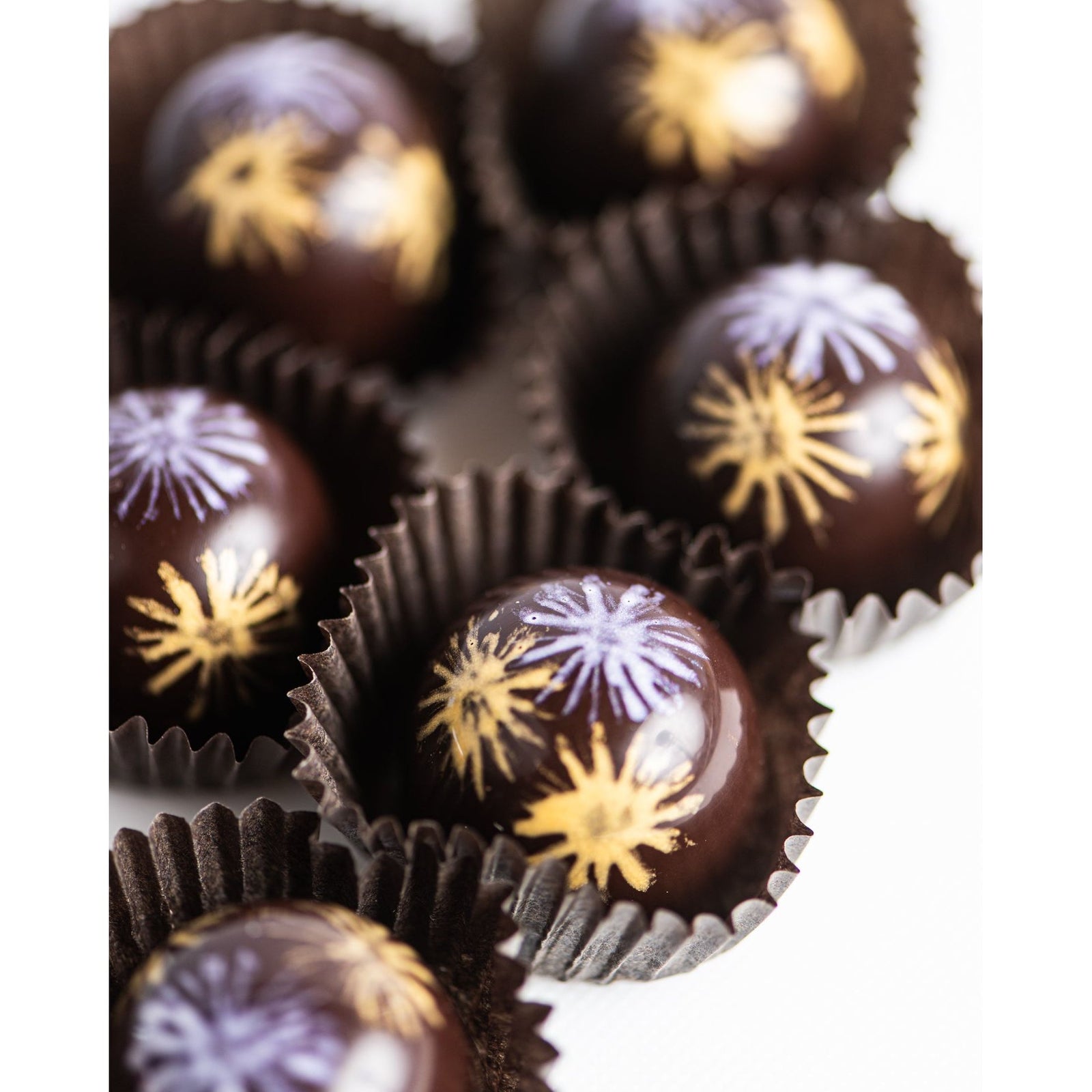 Close-up of glossy Champagne Bon Bons by Bixby Chocolates in brown paper cups, topped with hand-painted gold and purple floral or starburst designs.