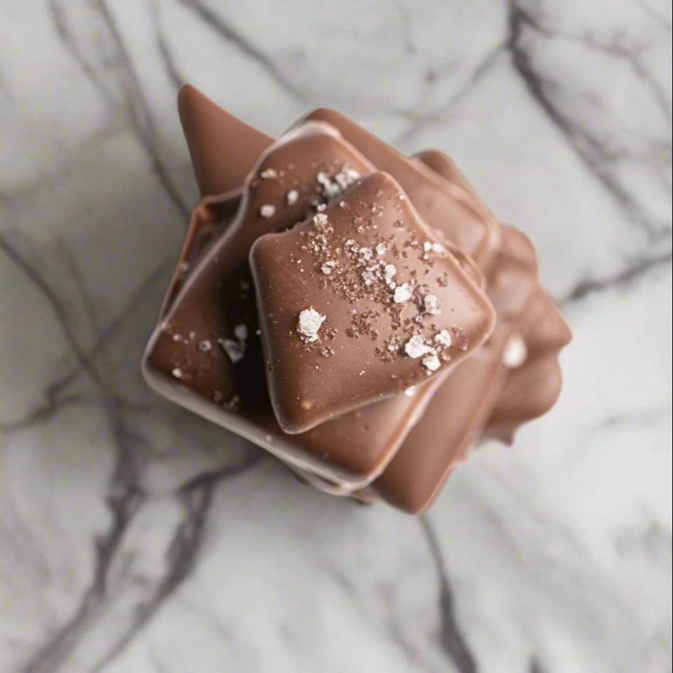 A close-up of Bixby Chocolates Milk Chocolate Toffee squares drizzled lightly and topped with sprinkled salt, arranged on a white marble surface.